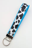 Handmade Holy Cow! key fob featuring a black and white cow spot print on a satin ribbon, shown on a turquoise nylon base with metal key ring.