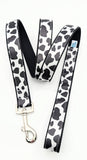 Handmade Holy Cow! leash featuring a black and white cow spot print on a satin ribbon, shown on a black nylon base with a silver metal snap.