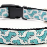 Close up of a dog collar featuring seafoam green cartoon T-Rex dinosaurs on a white ribbon, sewn onto durable [e.g., black] nylon webbing.